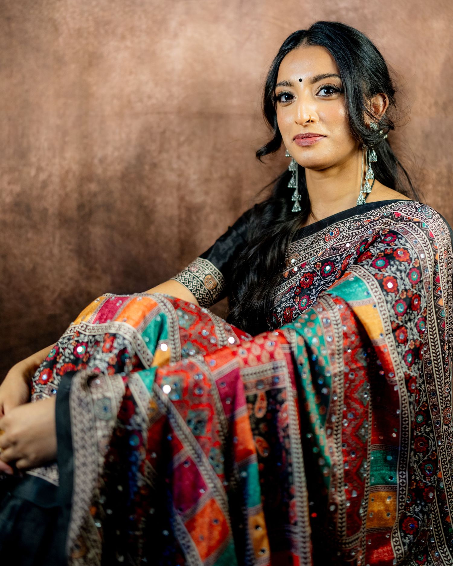 Woman in ornate black and multicolor embroidered saree with jewelry poses gracefully against brown backdrop.