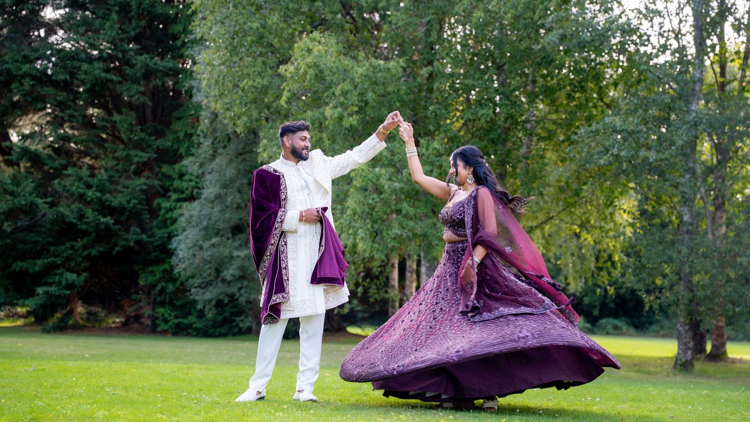 Indian bride spinning in purple embroidered lehenga while groom holds her hand on lush green lawn.