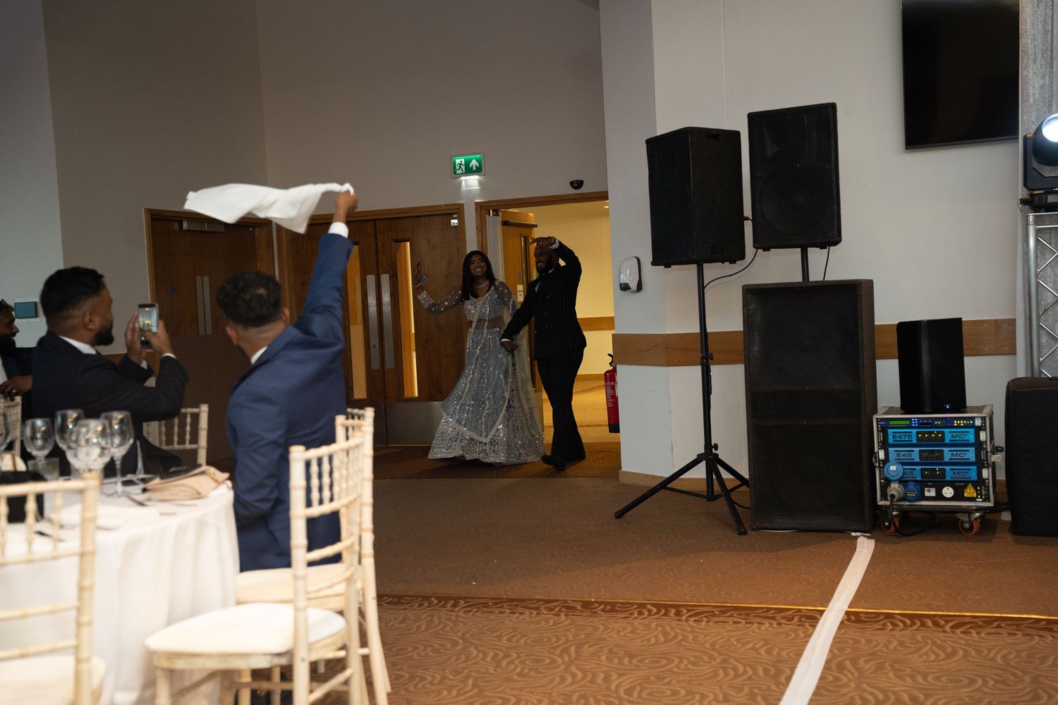 Groom waves white cloth as bride in blue lehenga enters wedding reception hall with DJ setup nearby.