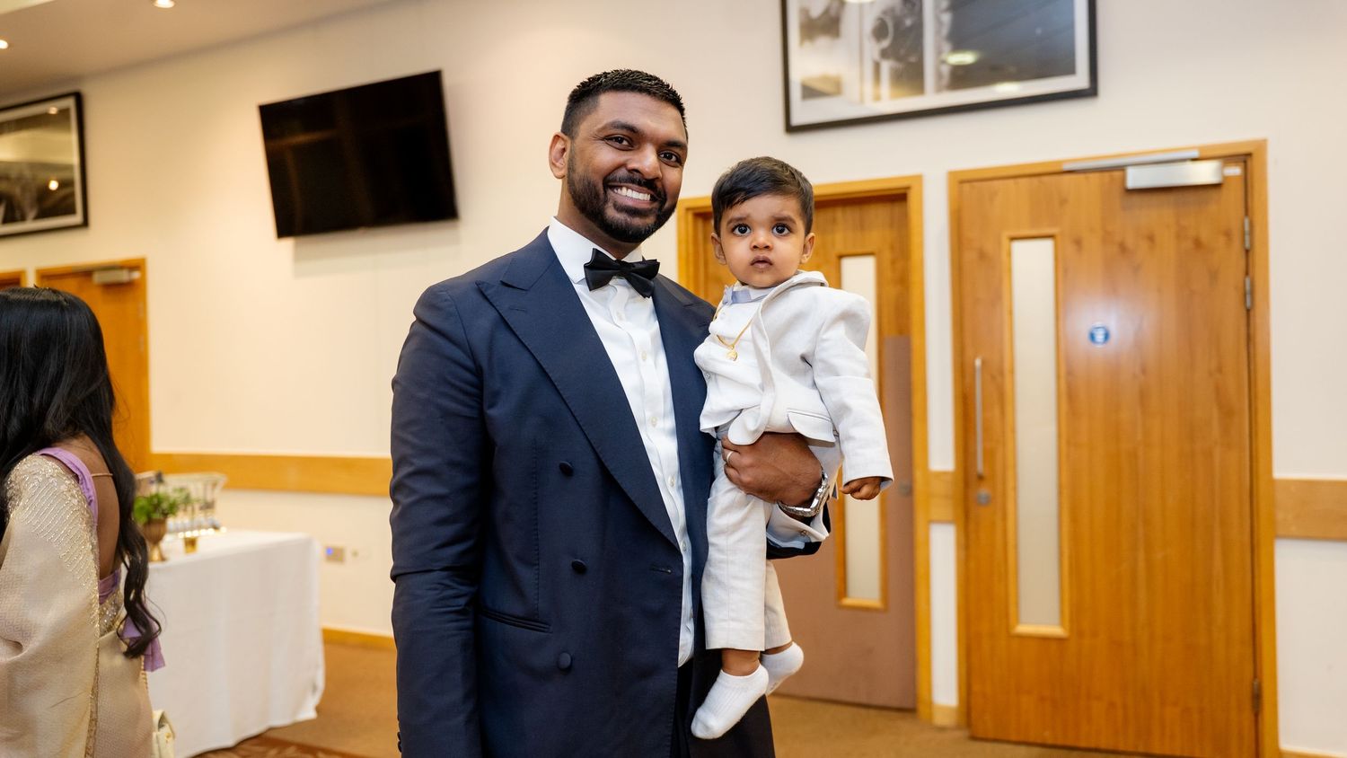 Smartly dressed man in navy tuxedo holding a baby in white outfit at a formal celebration.