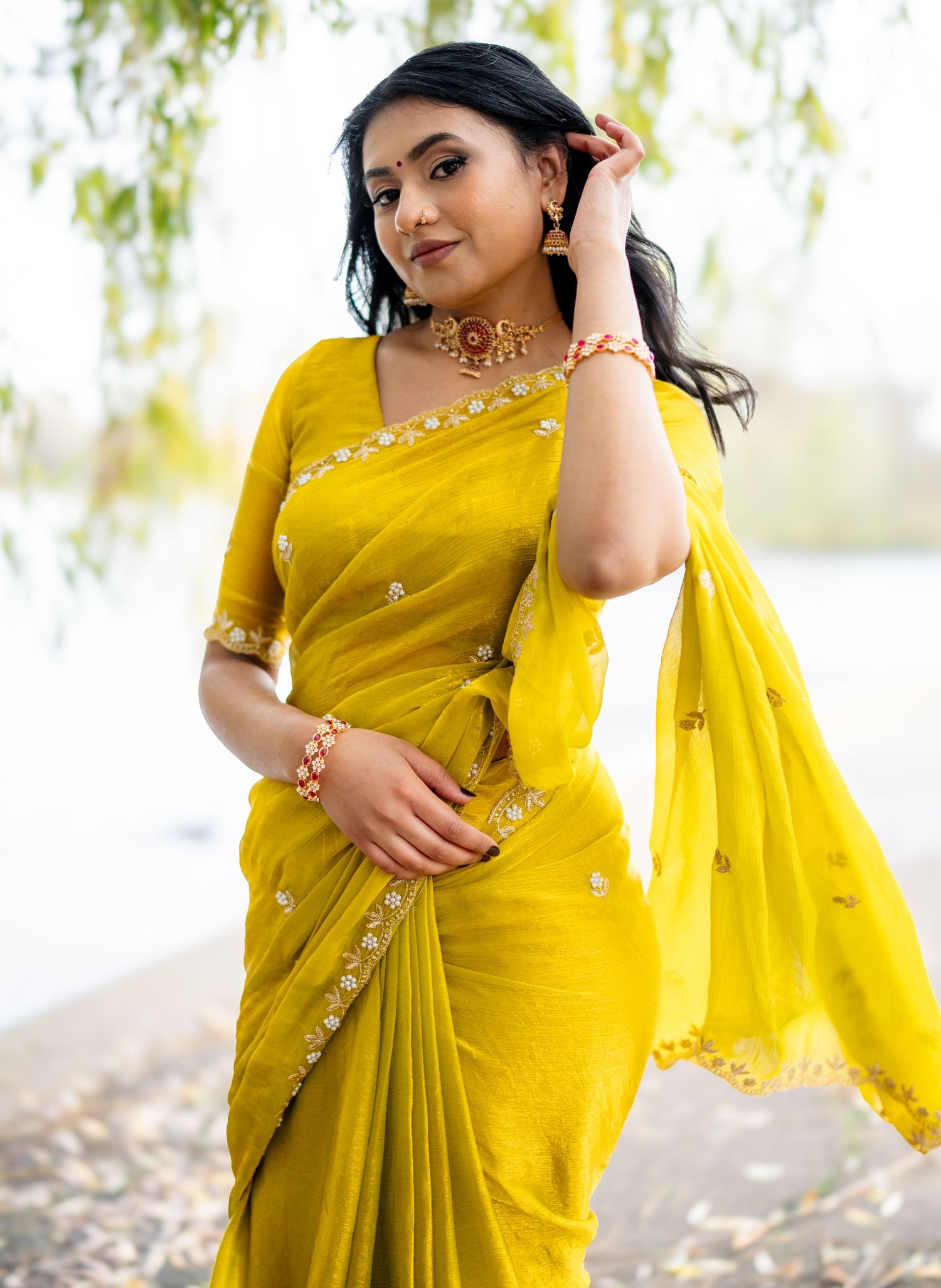 Beautiful woman in yellow embroidered saree with gold jewelry by a lake.