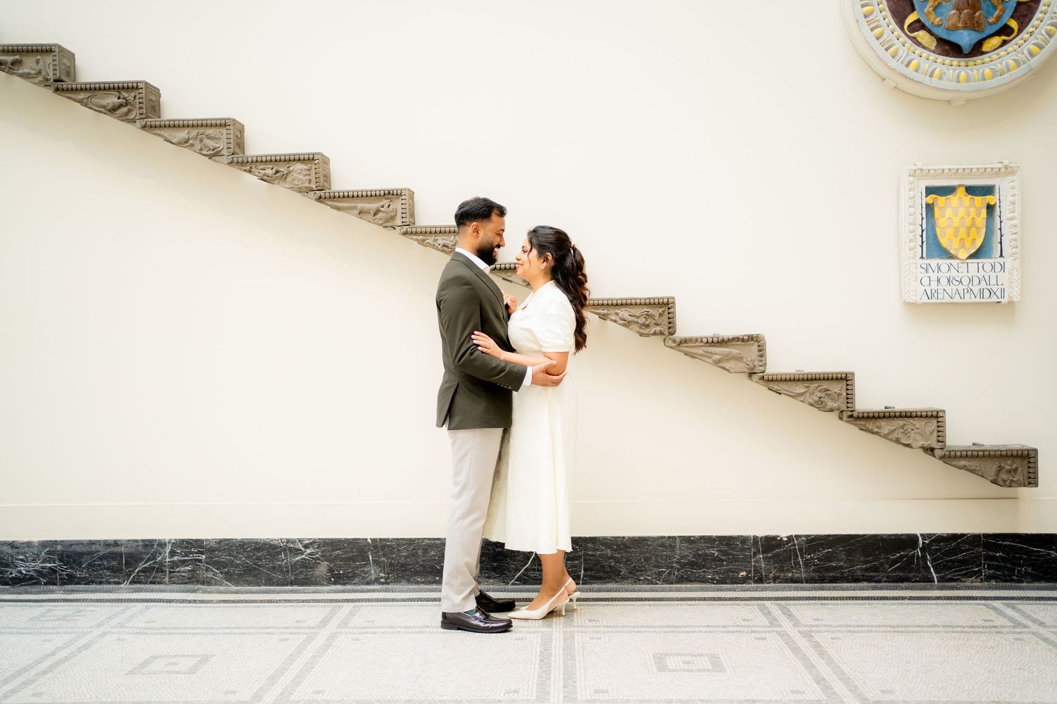 Couple embracing romantically near marble staircase in elegant building with decorative crest.