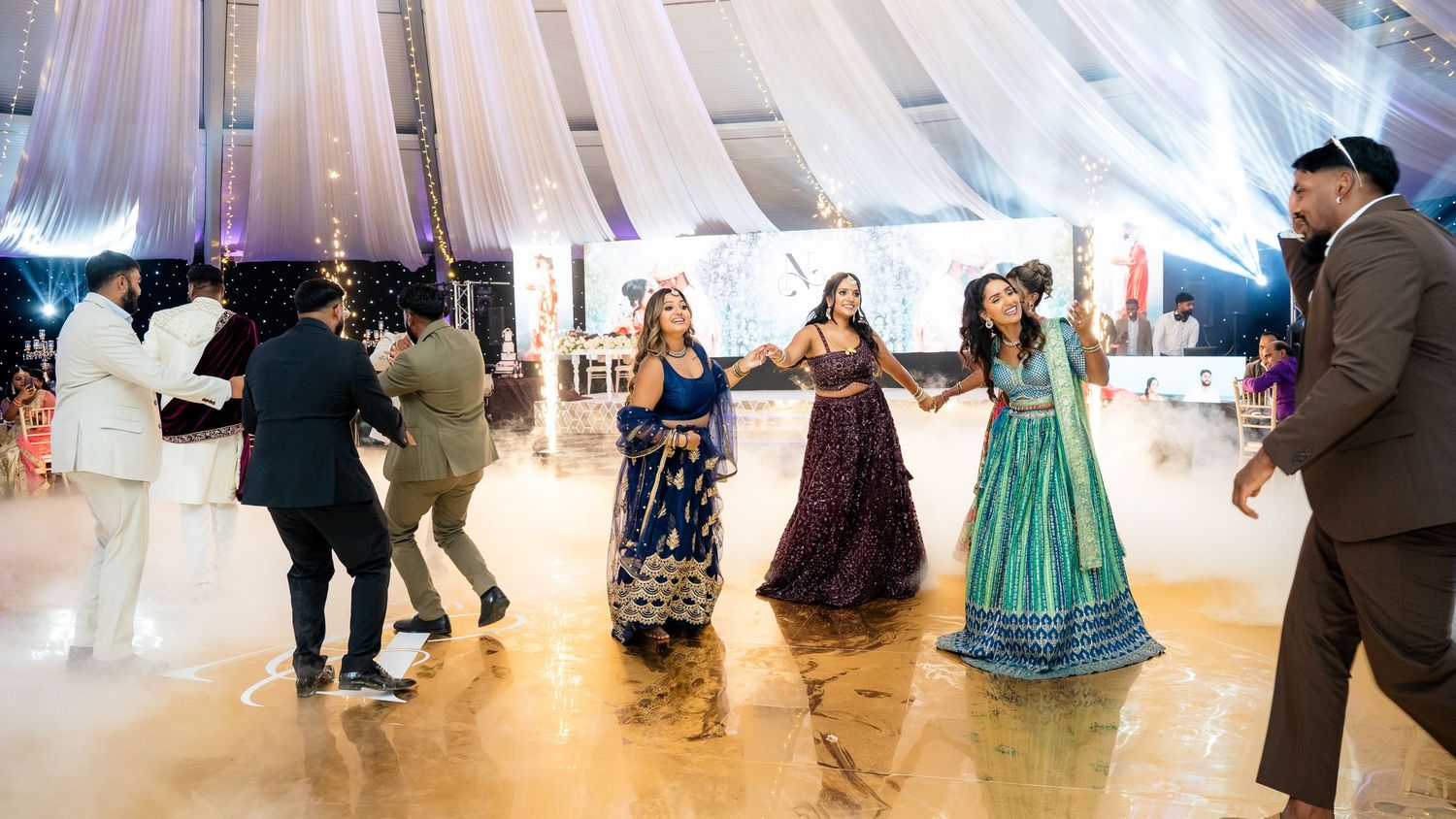 Guests in colorful traditional Indian attire dancing joyfully on the dance floor at a wedding reception.