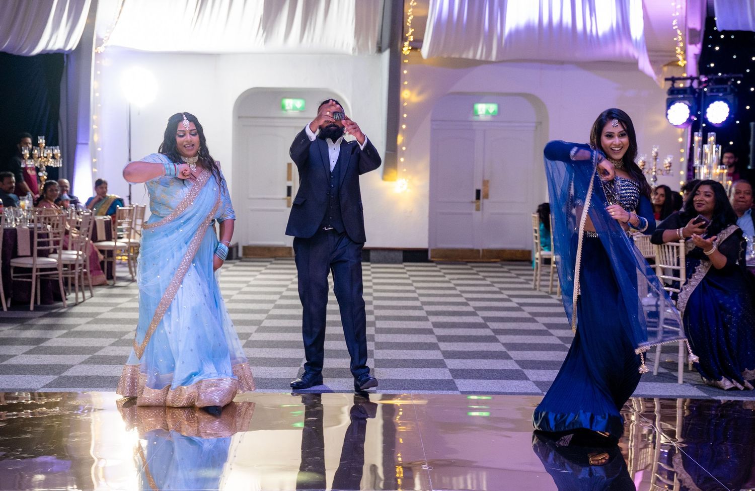 Three guests dancing joyfully in traditional Indian attire on a checkered ballroom dance floor.