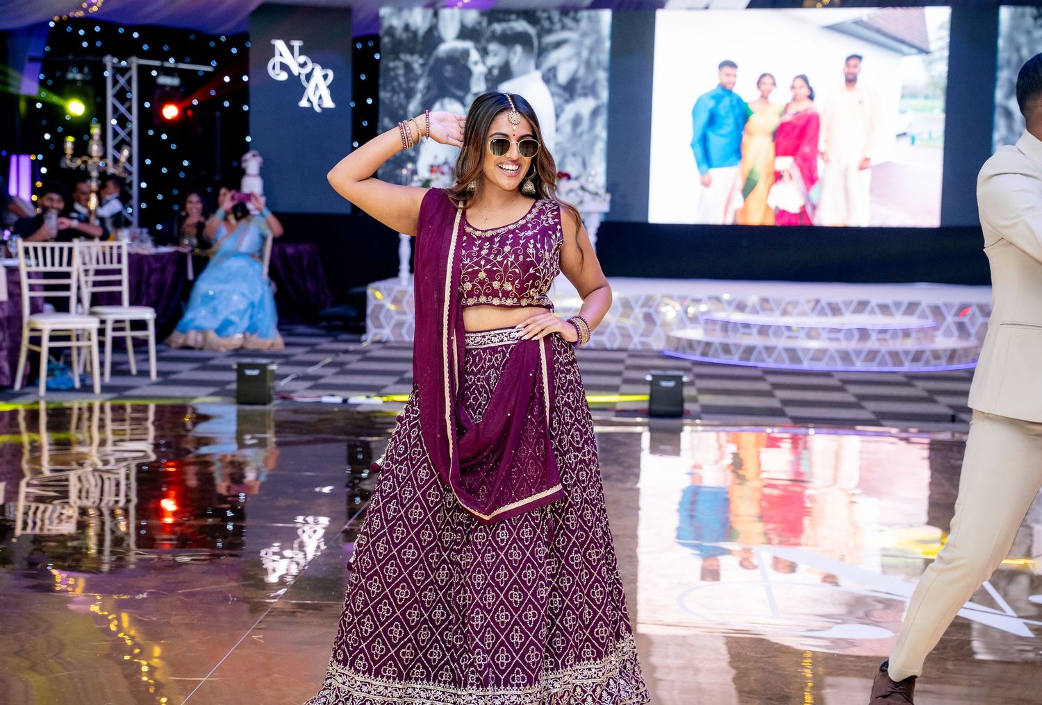 Woman in purple embroidered lehenga performing at wedding reception with stage and guests in background.