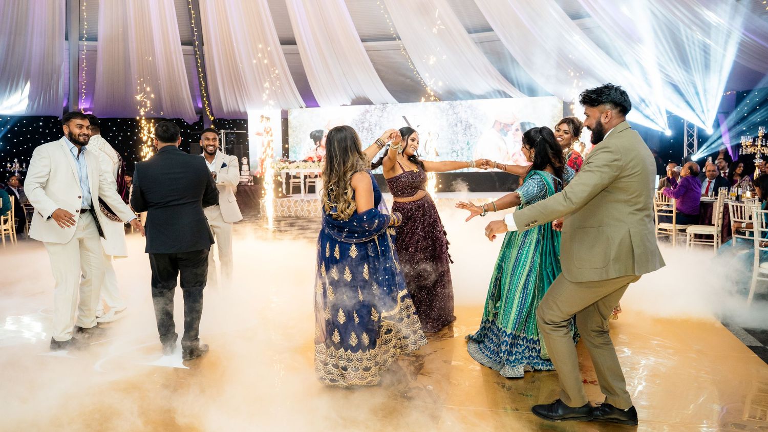 Guests in vibrant traditional Indian attire dance joyfully on a foggy white dance floor at an elegant wedding reception.