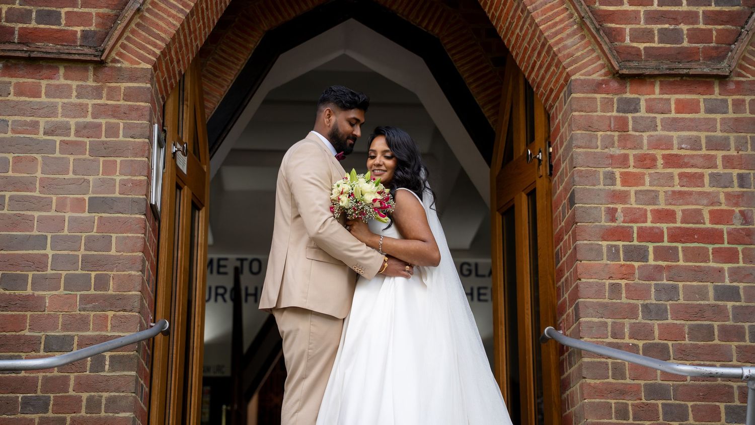 Couple in wedding attire embracing outside a brick church, bride holding a bouquet in a white gown.