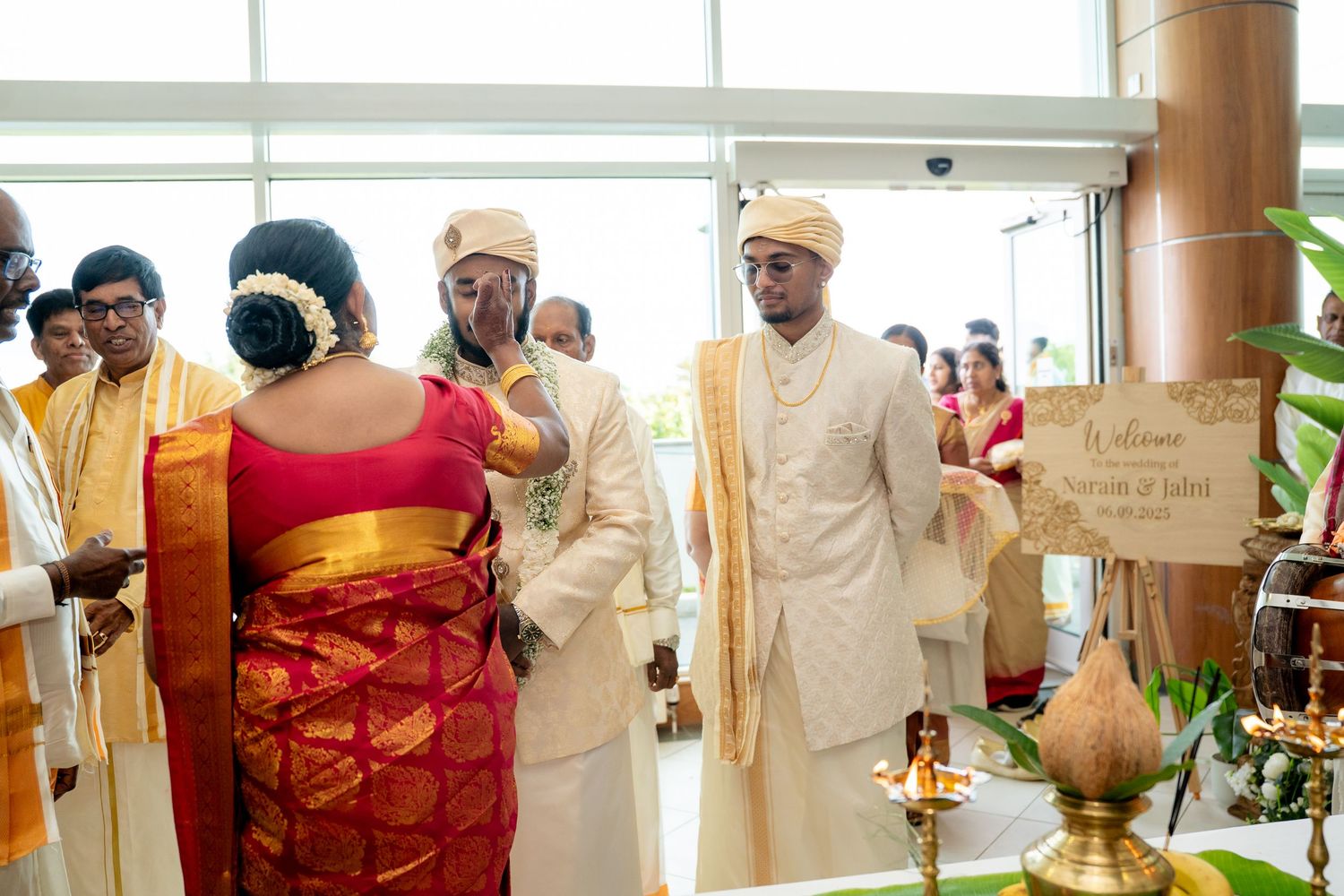 Bride in red and gold saree meeting groom in cream sherwani at a decorated traditional Hindu wedding venue entrance.