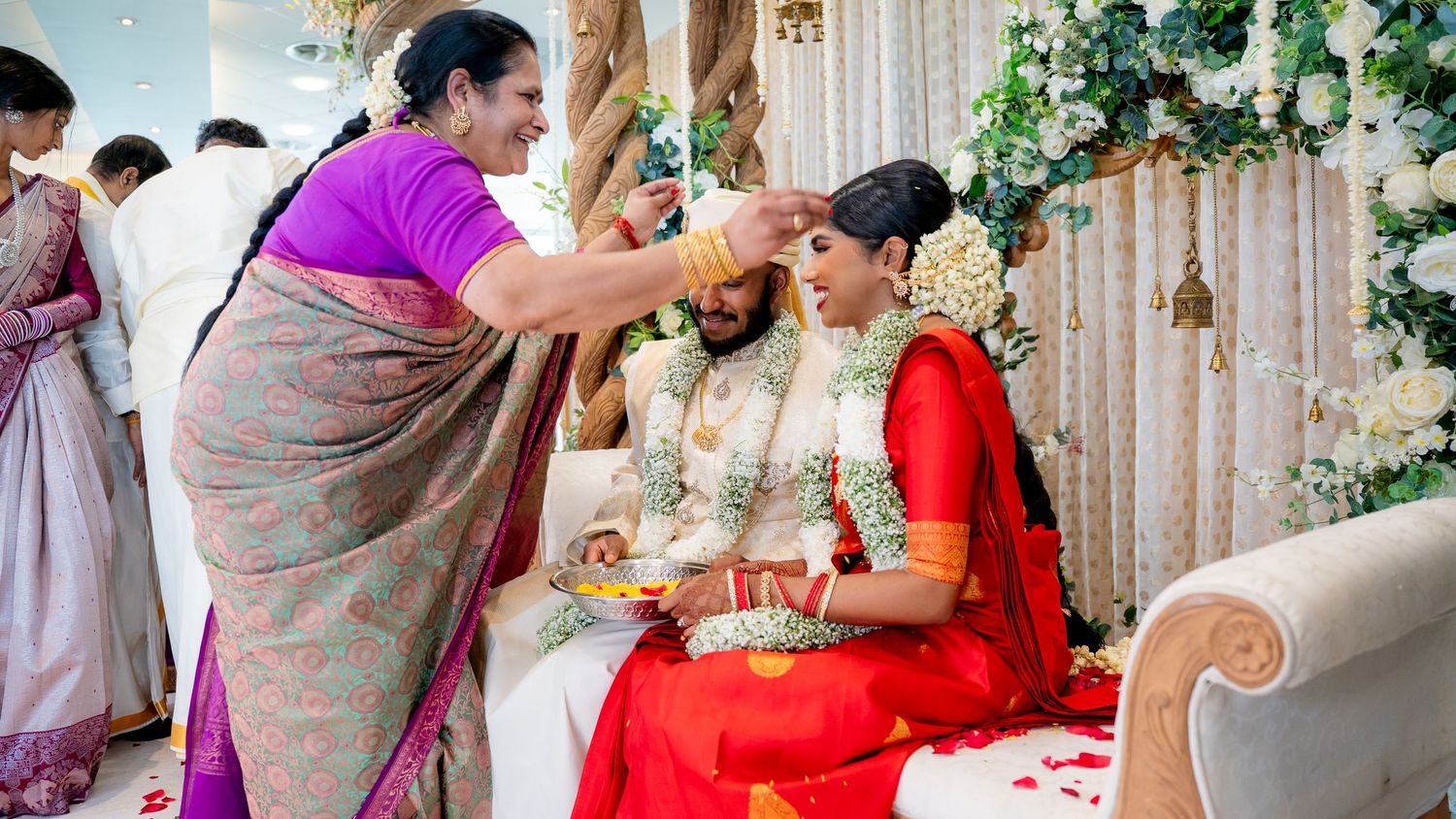 Elder woman applying tilak to Hindu bride and groom seated at decorated wedding mandap ceremony.