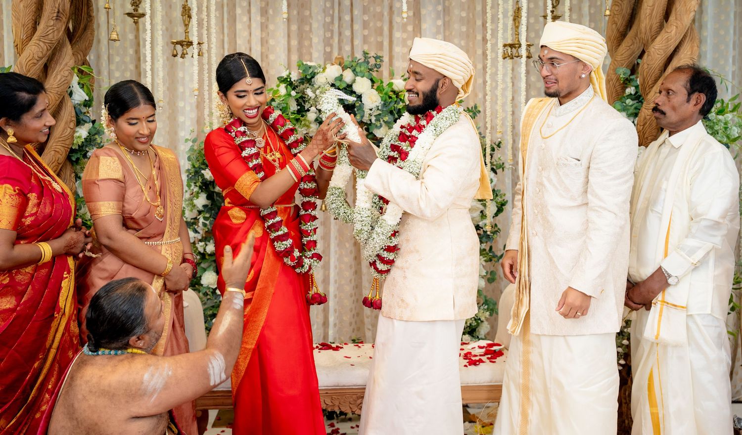 Smiling South Indian wedding couple exchanges garlands on floral decorated stage with guests in attendance.
