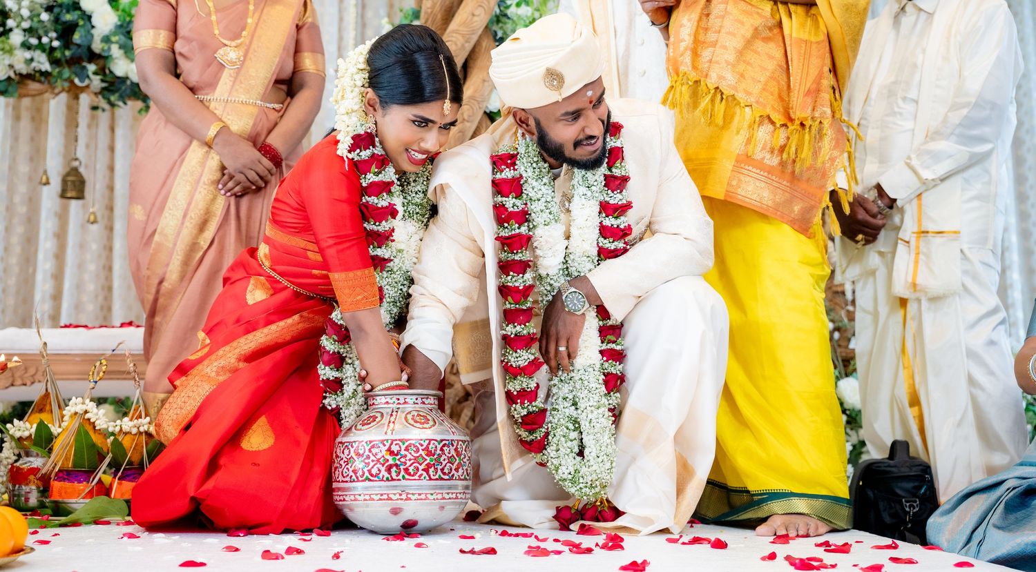 Indian couple performing traditional Hindu wedding ceremony with flower garlands and red petals at colorful mandap.