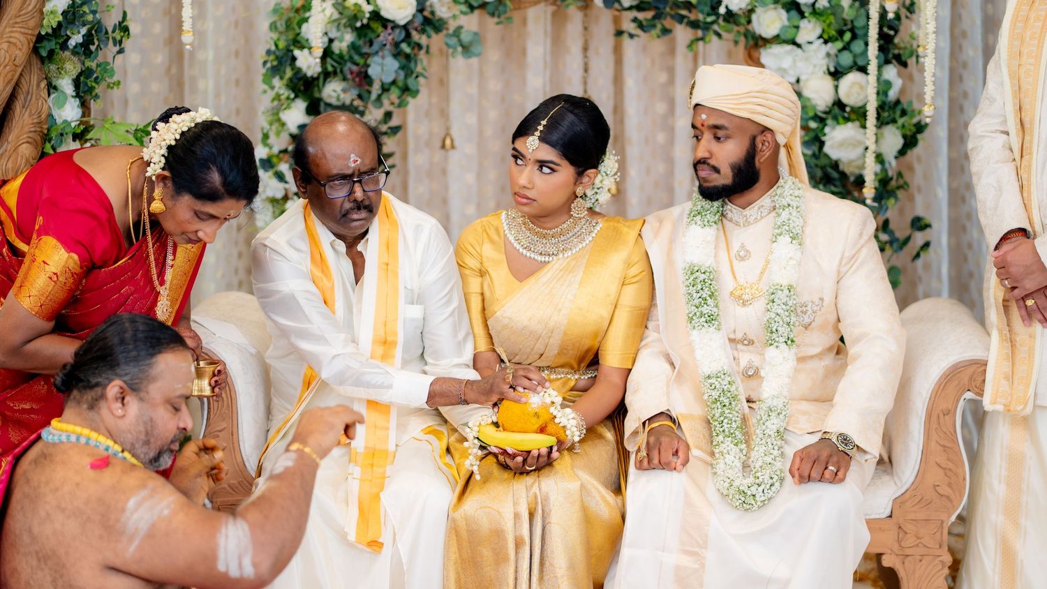 Traditional South Indian wedding ceremony with bride in gold saree and groom in white sherwani seated at mandap.