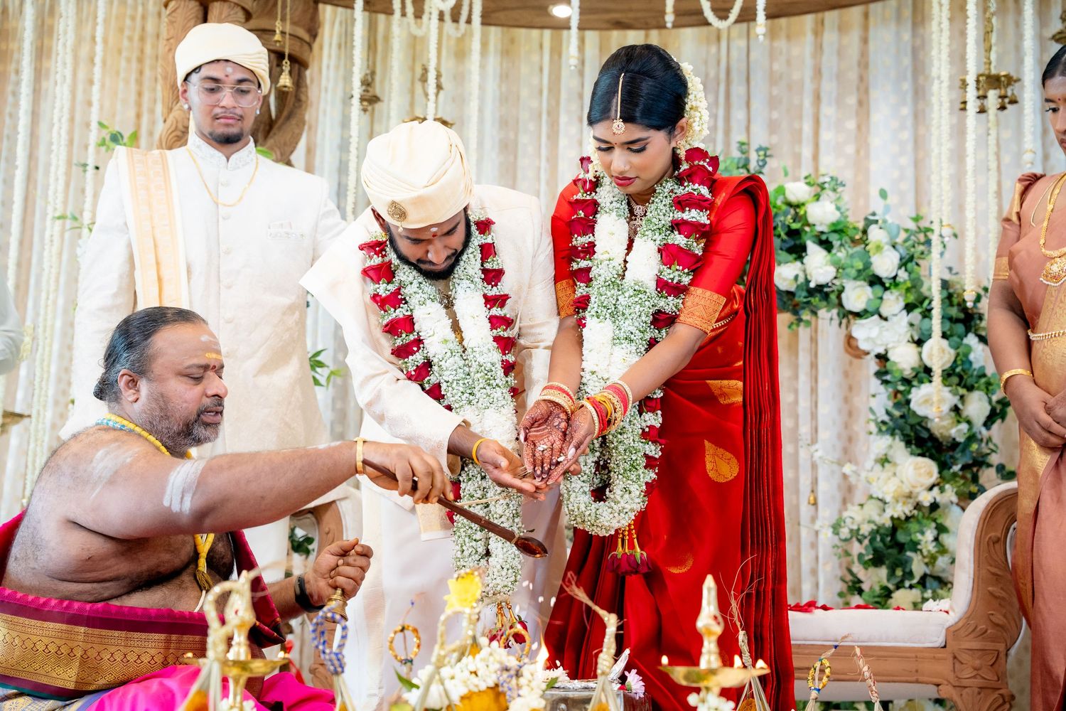 Hindu bride and groom conduct traditional wedding ceremony with priest at flower-adorned altar.