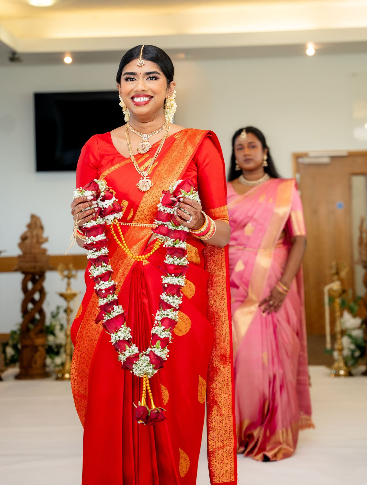 Radiant South Indian bride in red saree smiles while holding white jasmine wedding garlands.