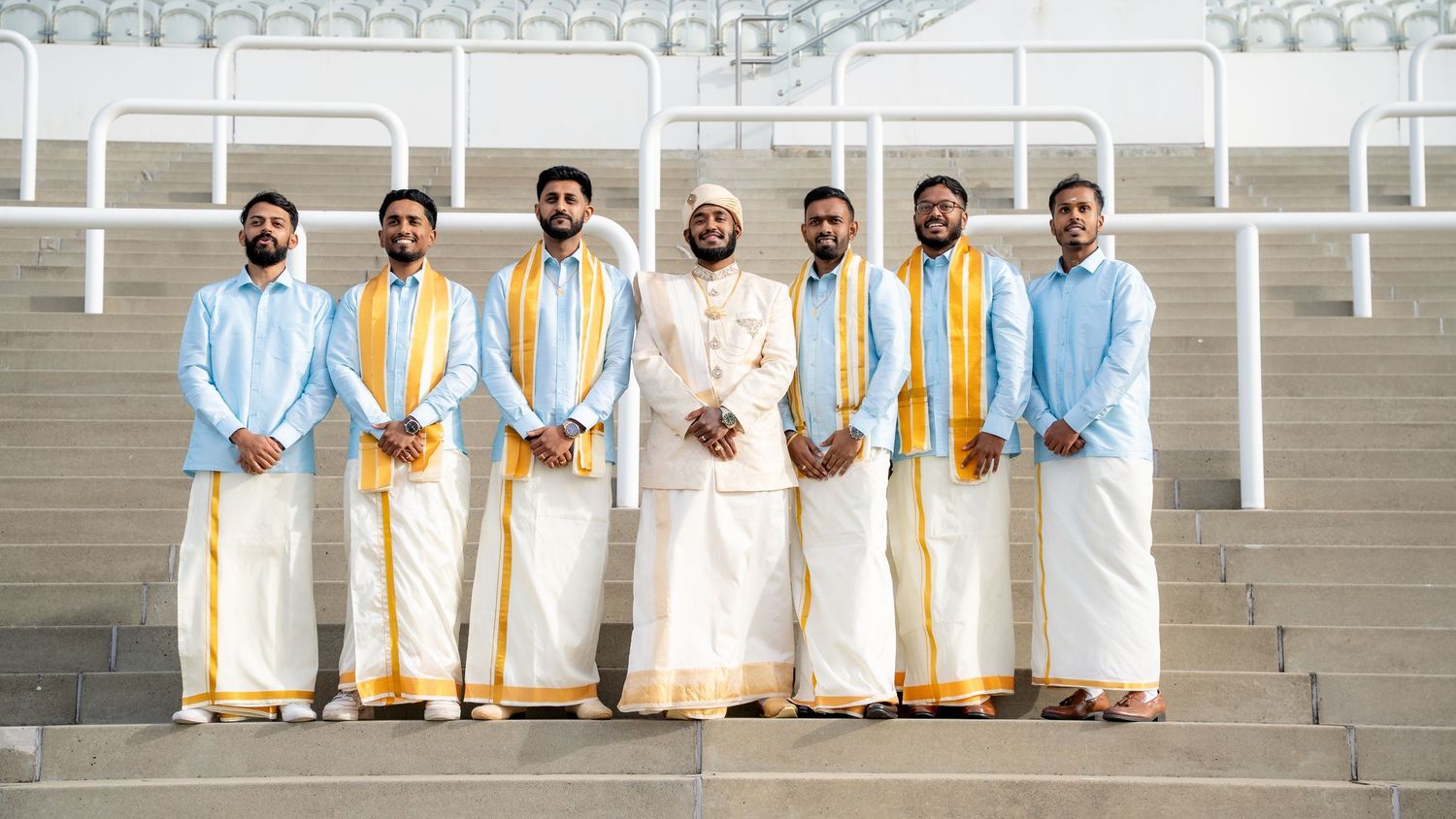 Wedding party in matching traditional South Asian attire posing together on outdoor stadium steps.
