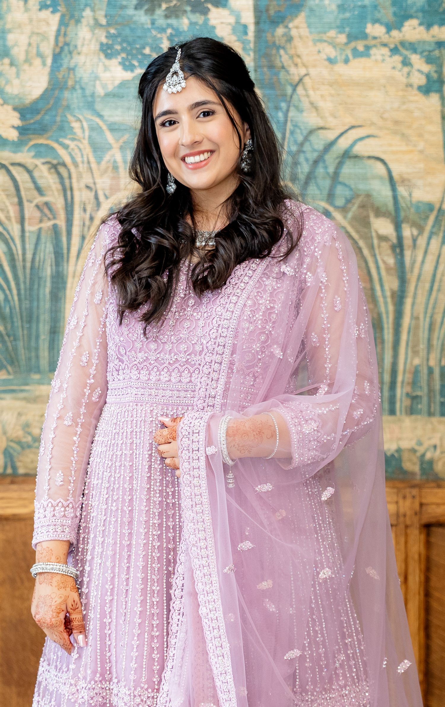 Young woman in lavender embroidered anarkali suit with dupatta and maang tikka, smiling at a formal event.