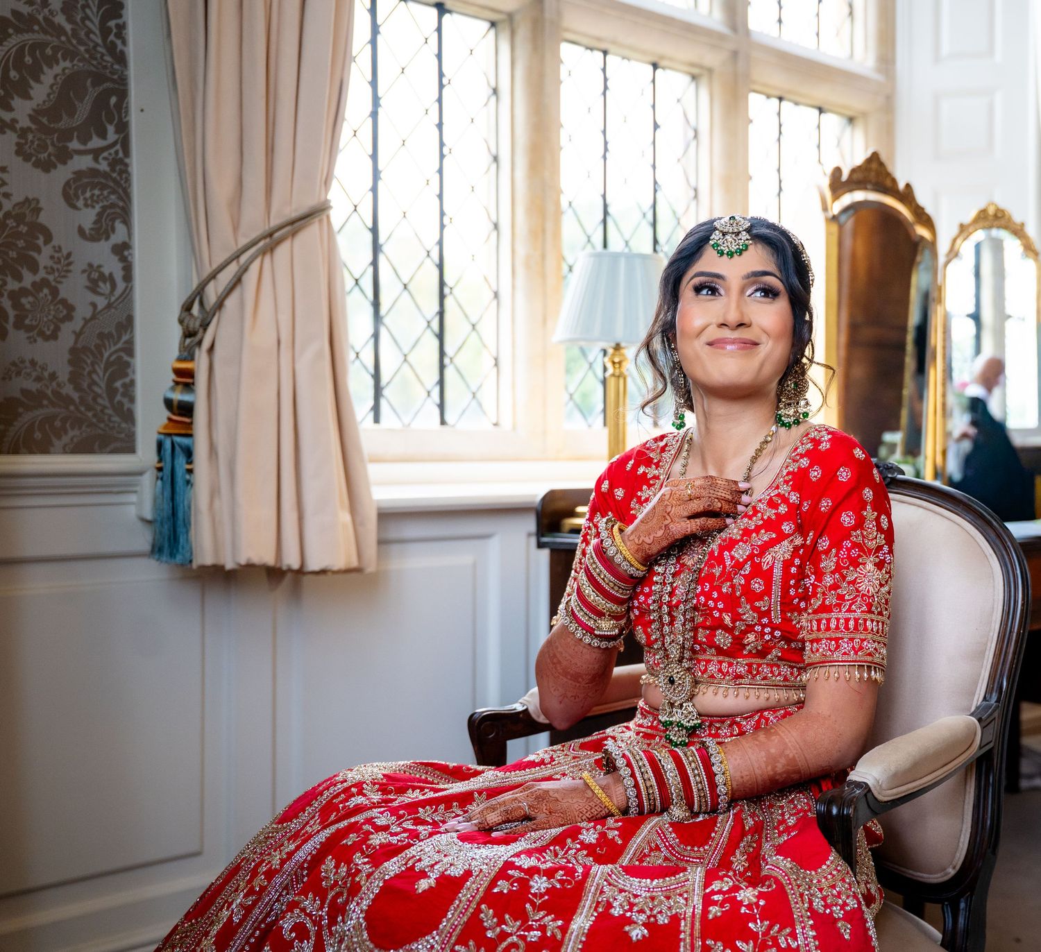Indian bride in red embroidered lehenga seated by window with bridal jewelry and bangles.