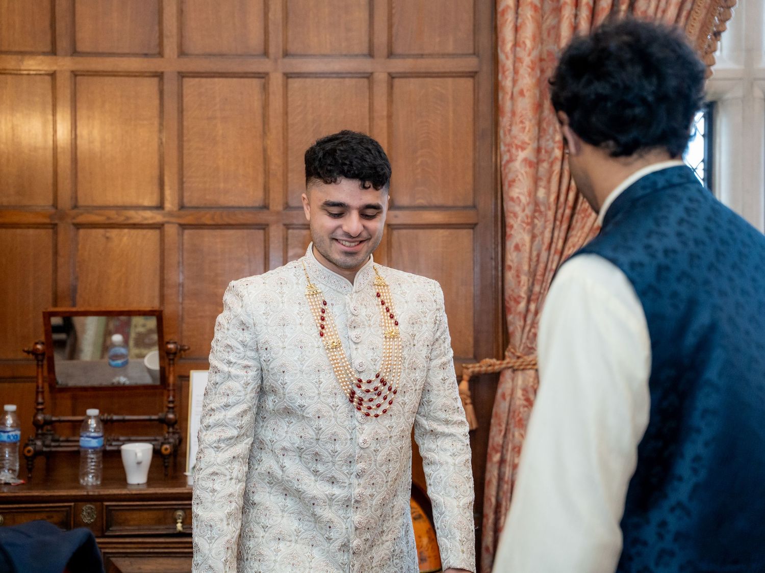 Groom in ornate white sherwani with floral garland smiles during Indian wedding preparations.