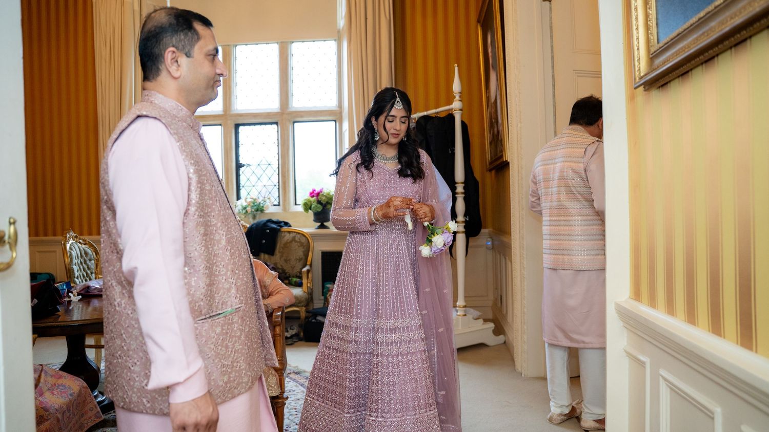 Bride and groom in pink traditional Indian wedding attire prepare in an elegant room.