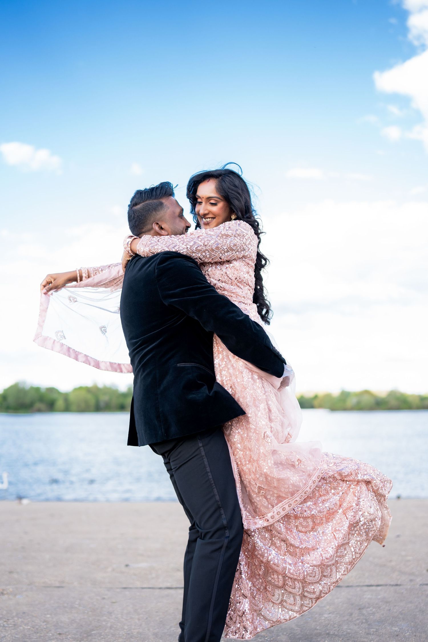 Groom lifts bride in pink lehenga by waterfront during romantic outdoor engagement photo session.