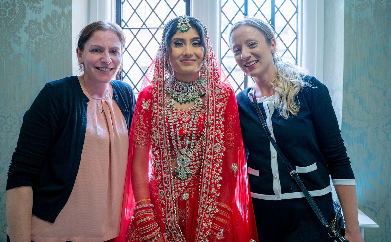 Bride in red Indian bridal attire with jewelry poses with two women in formal wear indoors.
