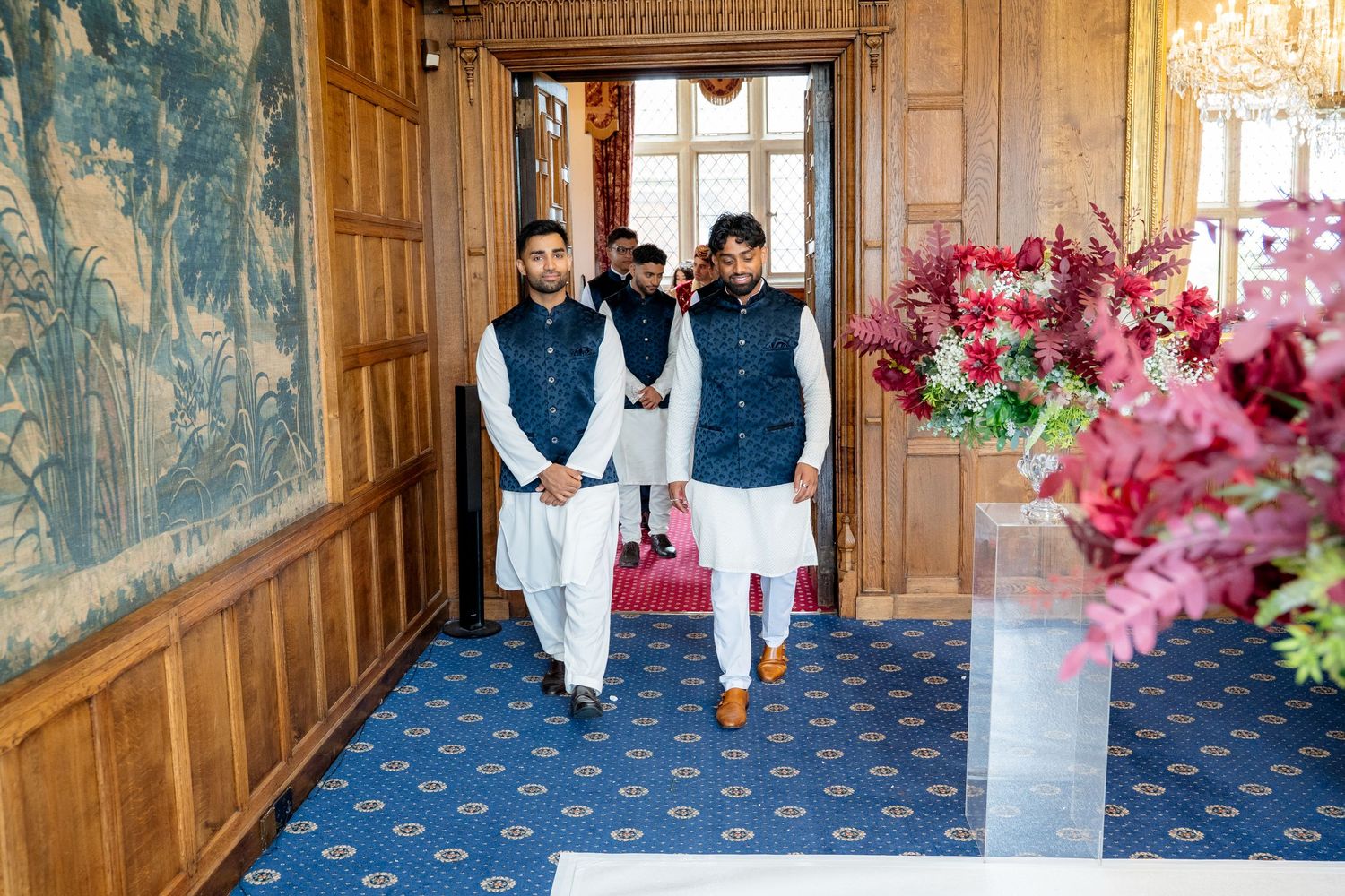 Men in traditional white and navy Indian attire walking through wood-paneled wedding venue.