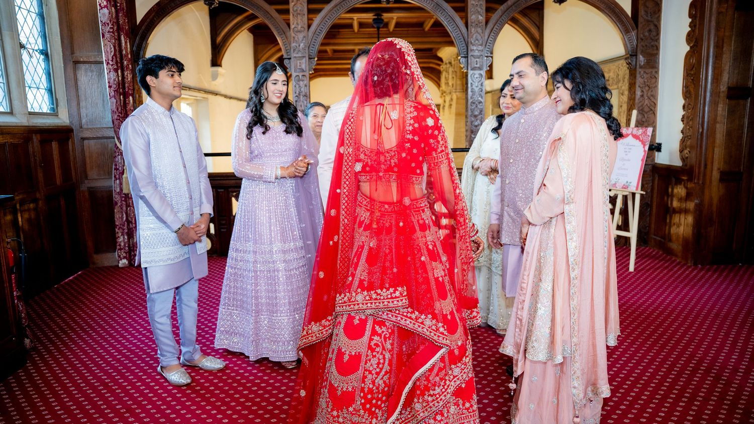 Indian bride in red lehenga greeted by family members in pink outfits inside elegant wedding venue.
