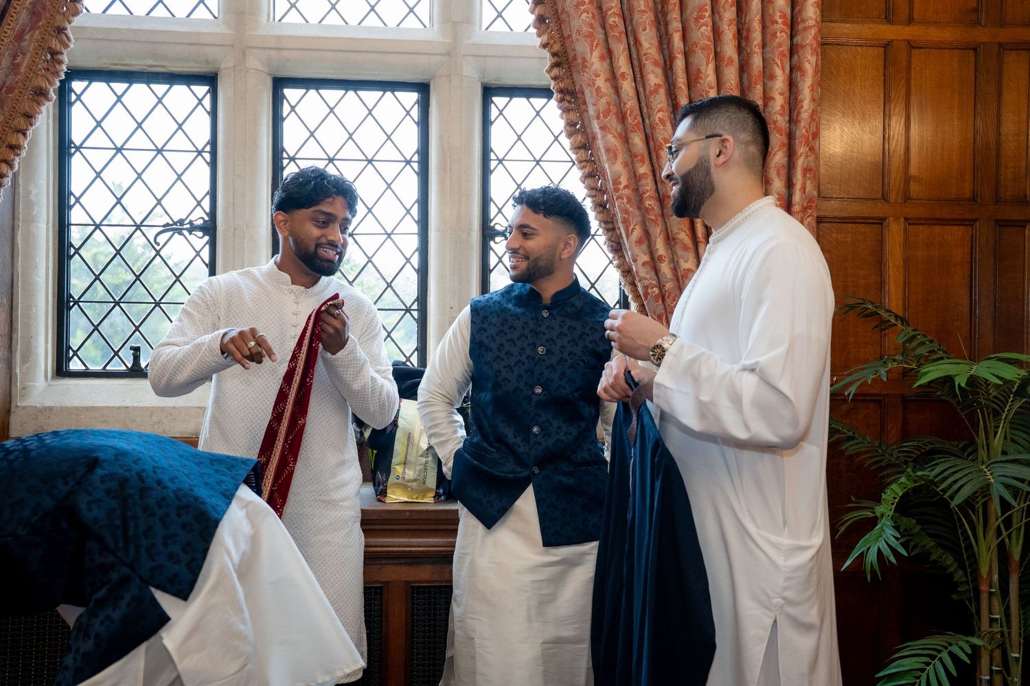 Three men in traditional South Asian attire chat indoors, one holding a red tie, near elegant windows.