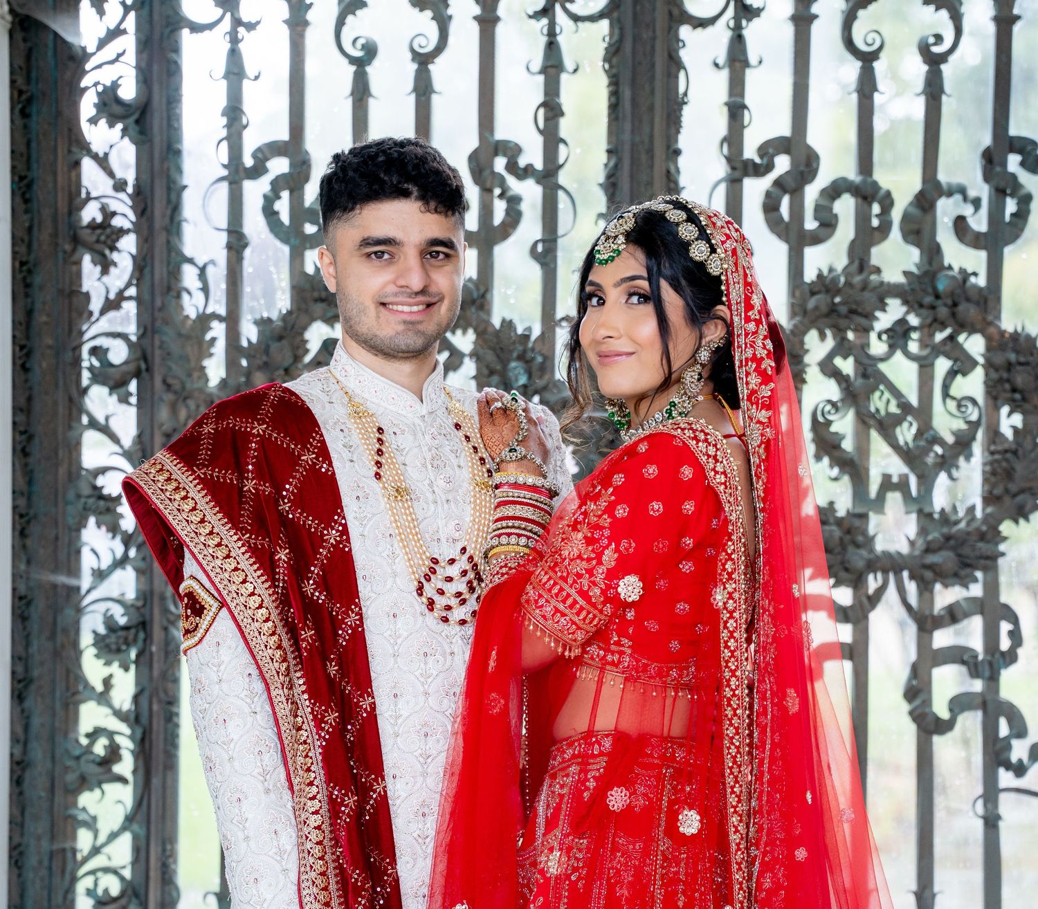 Couple in traditional red and white South Asian wedding attire posing before ornate iron gates.