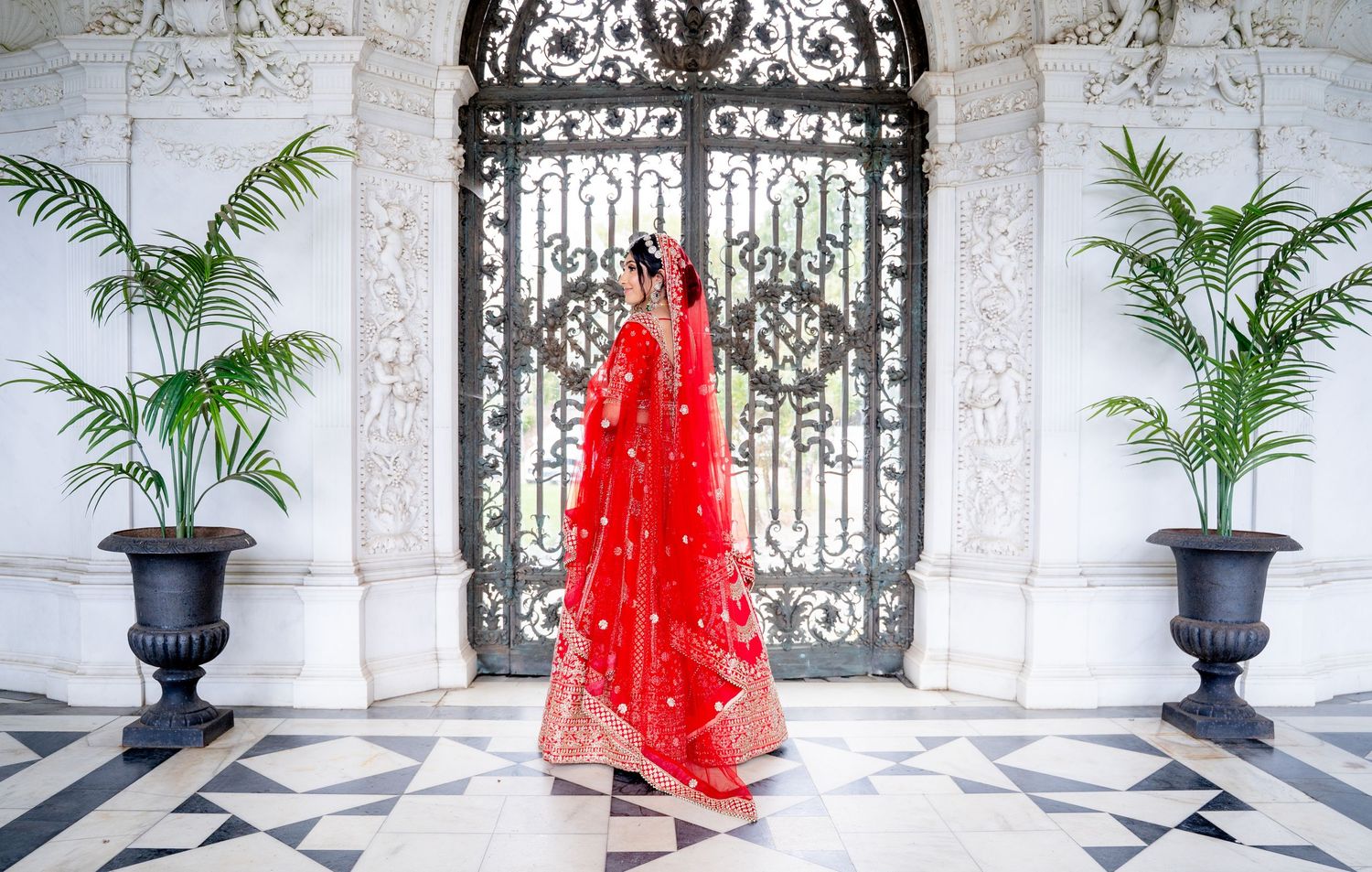 A bride in a stunning red lehenga stands before ornate iron gates flanked by tropical plants on a marble floor.
