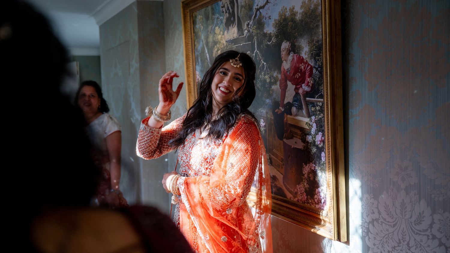 Indian bride in orange saree laughing joyfully during wedding celebration indoors with guests nearby.