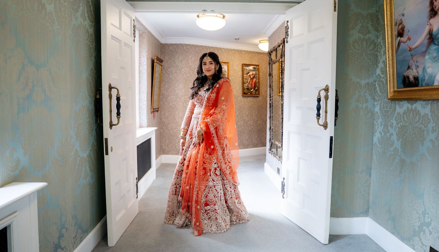 South Asian bride in embroidered orange lehenga poses gracefully in bright hotel corridor.