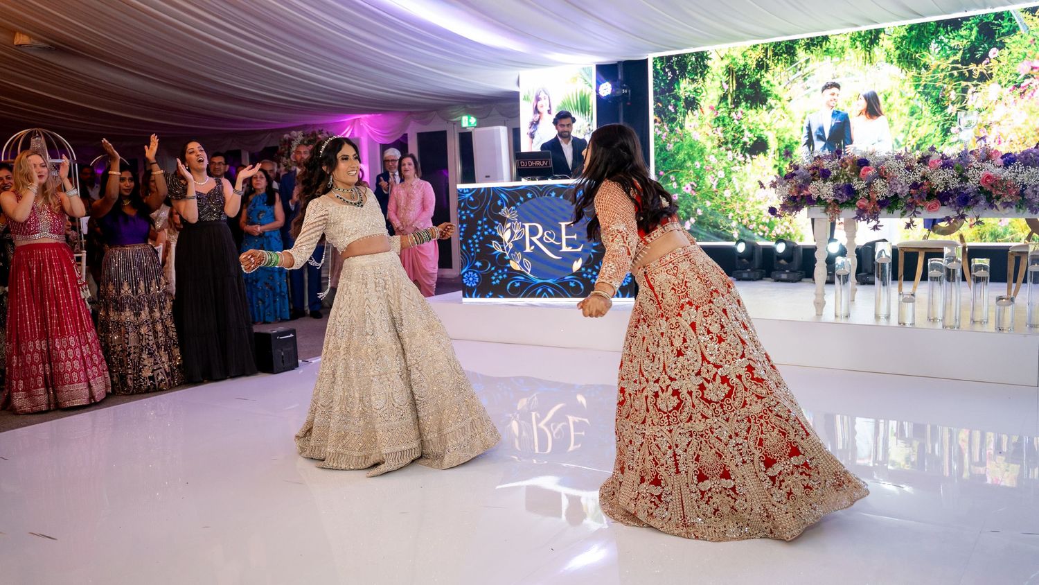 Women in ornate traditional lehengas dancing at an elegant indoor wedding reception with purple lighting and guests watching.