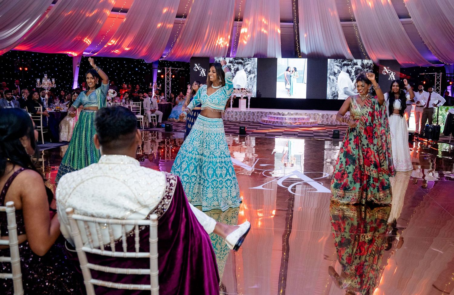 Women in traditional Indian attire dancing at a decorated wedding reception in a tent.