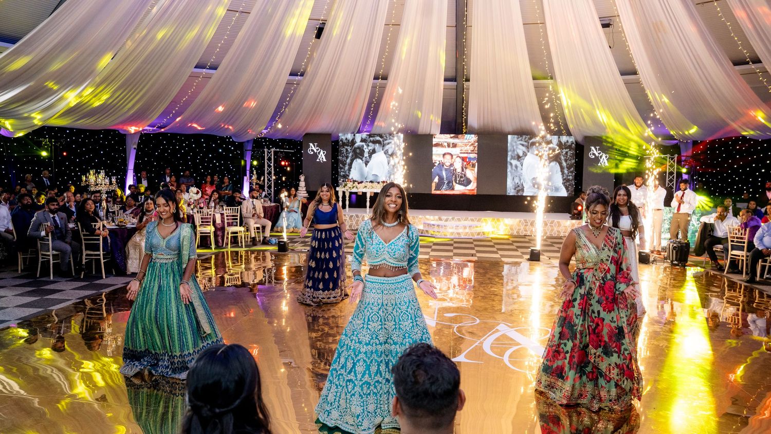 Elegant quinceañera celebration with young women in colorful formal gowns dancing on a decorated ballroom floor with dramatic lighting.