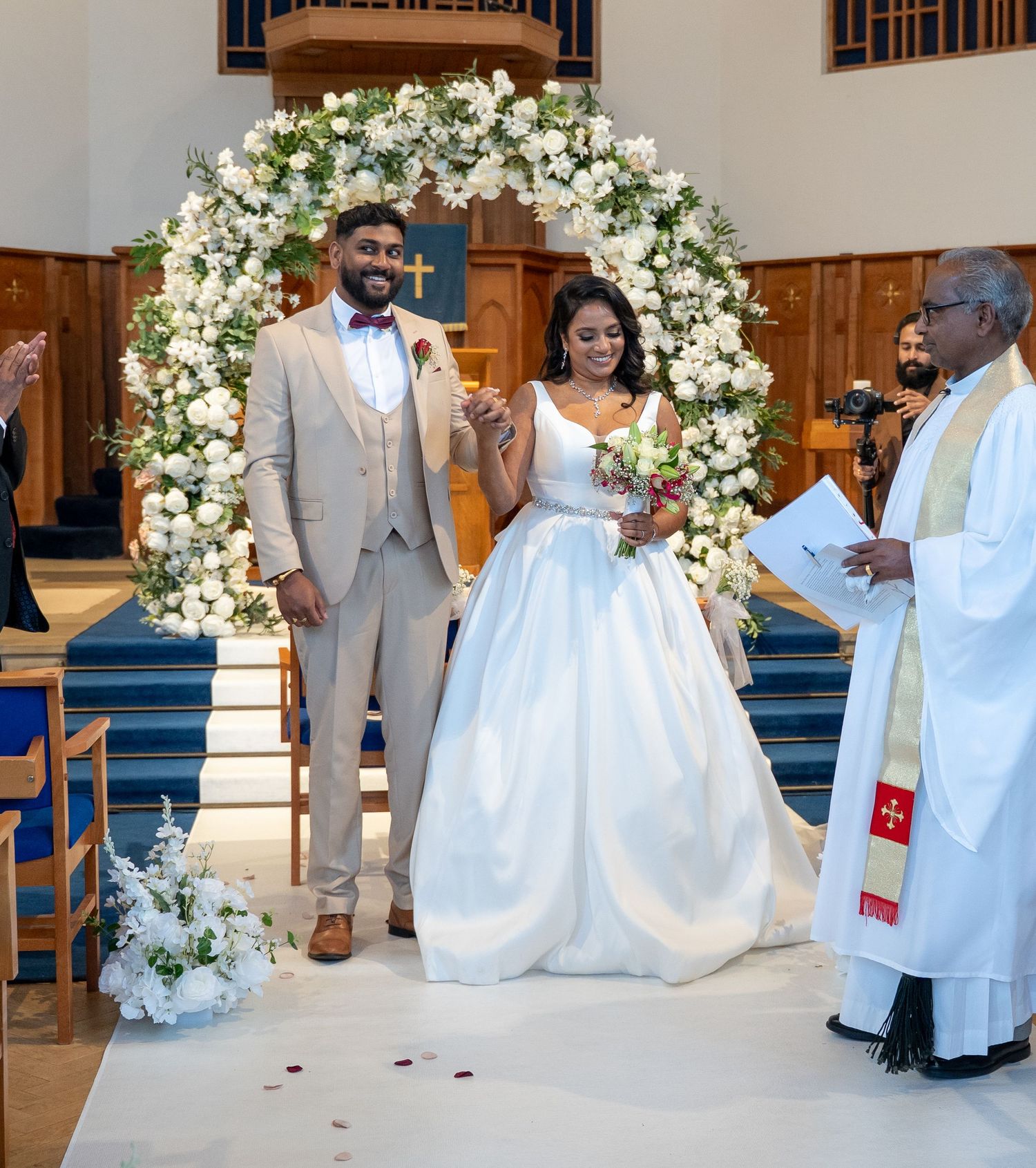 Joyful newlyweds stand before a floral arch at church altar as officiant reads from ceremony script.