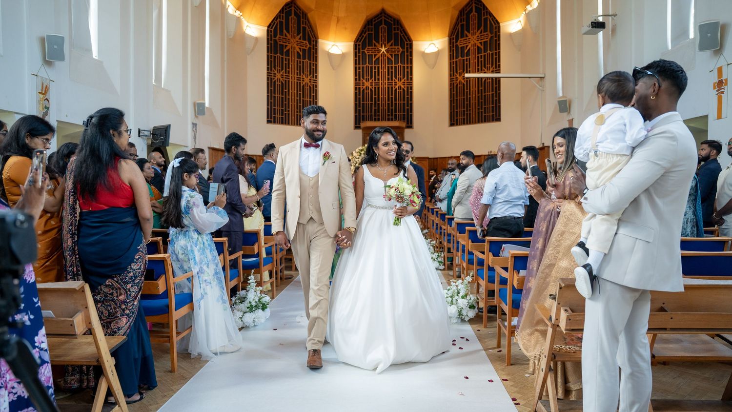 Bride and groom walk down church aisle during wedding ceremony, surrounded by guests in elegant attire.