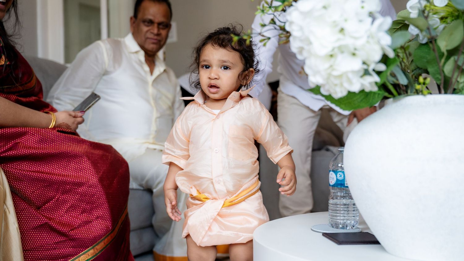 Cute baby in peach traditional Indian outfit standing near white floral arrangement at wedding event.