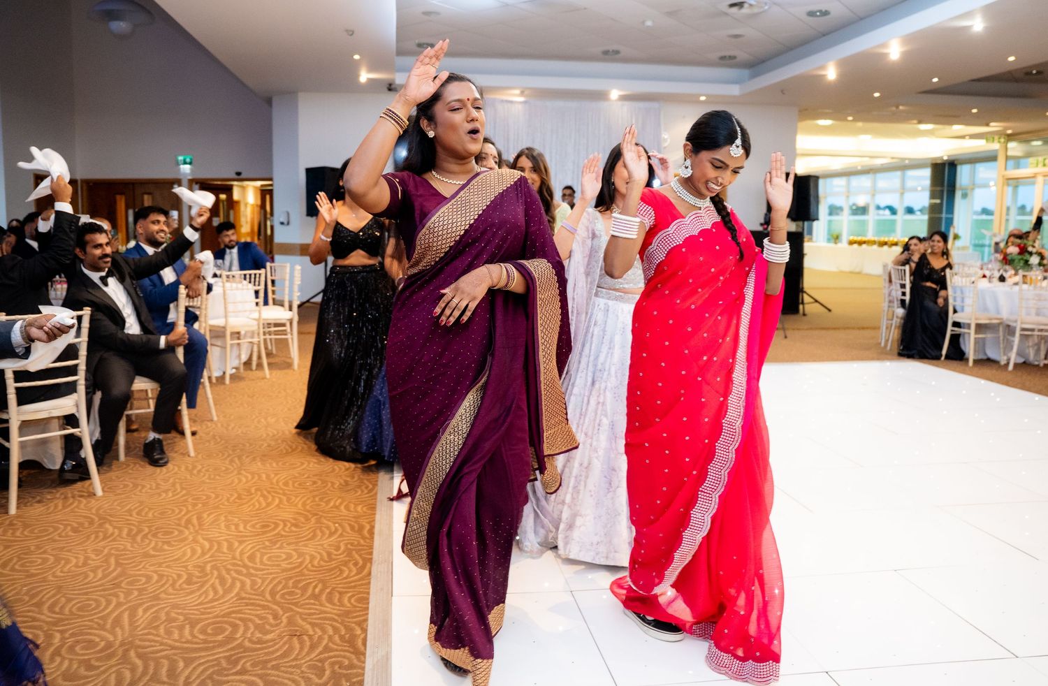 Women in sarees performing a traditional Indian dance on white dance floor at wedding.