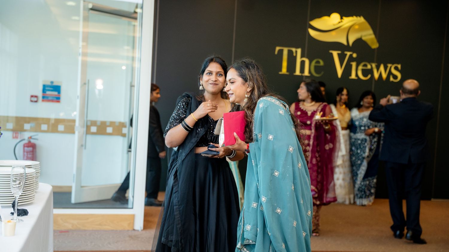 Two women in elegant sarees chatting near The Views venue entrance at a formal event.