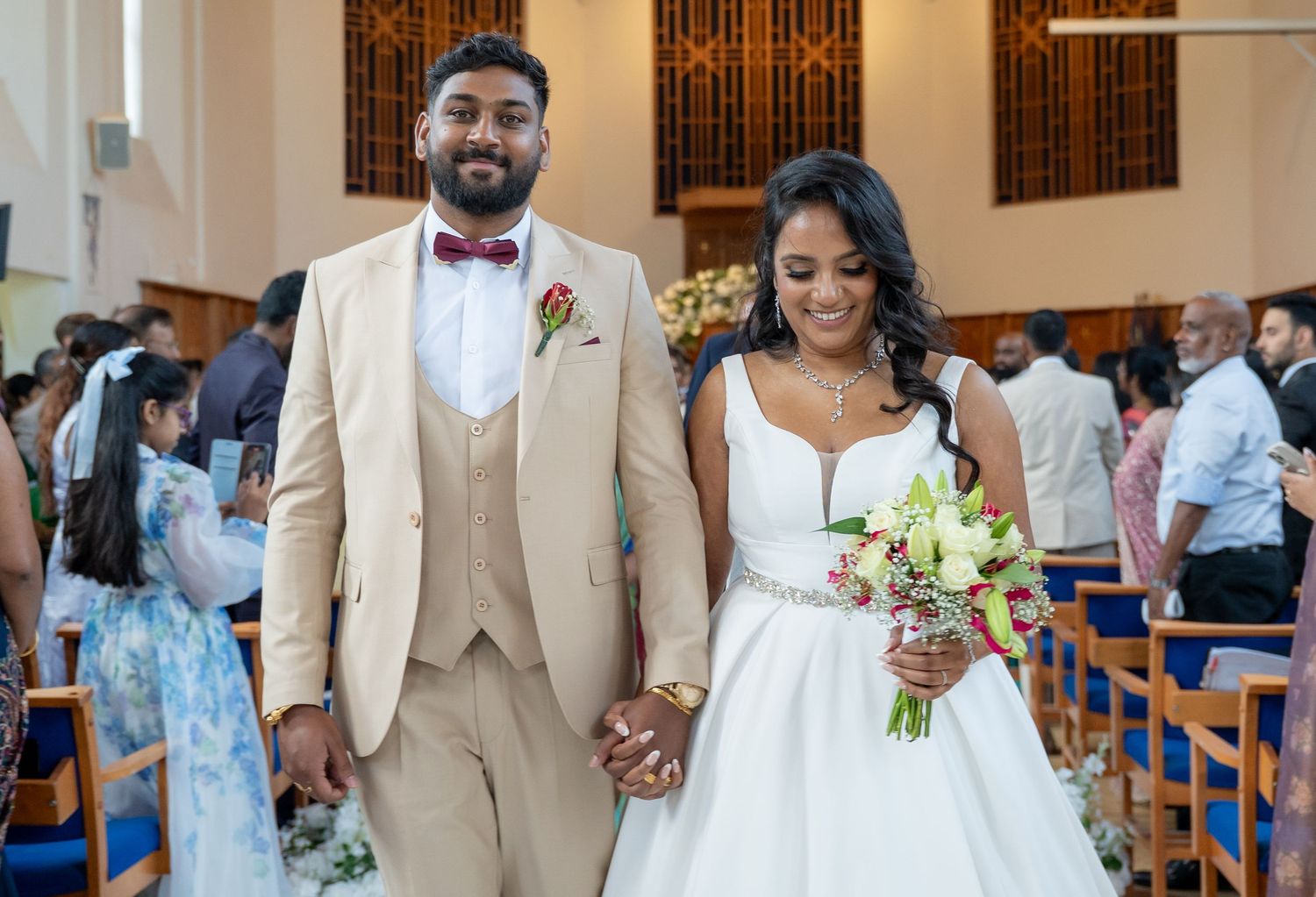 Bride and groom walking down the aisle together in a church wedding ceremony, groom in beige suit, bride in white gown.