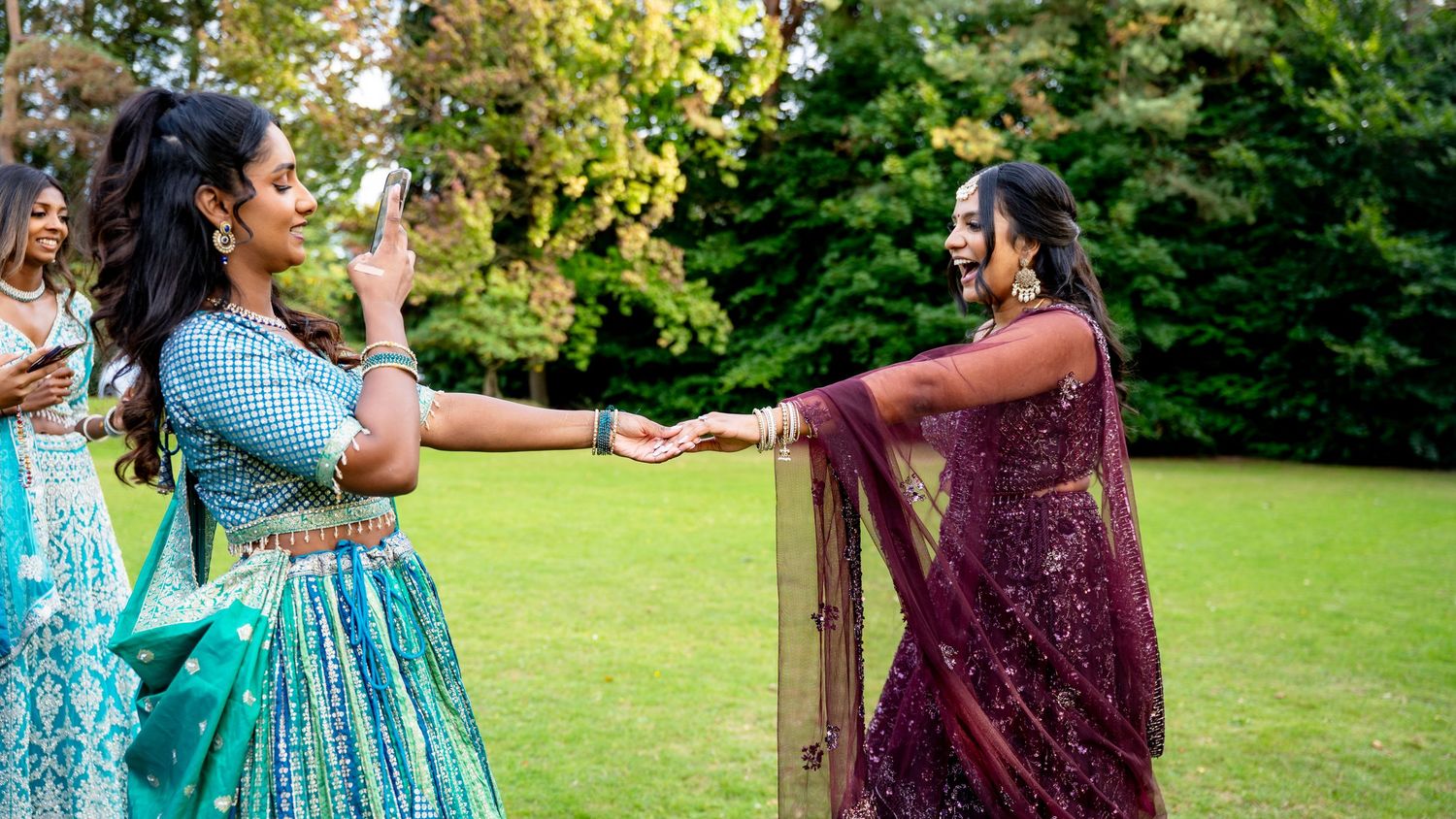 Two women in colorful Indian lehengas hold hands and dance together on a green lawn.