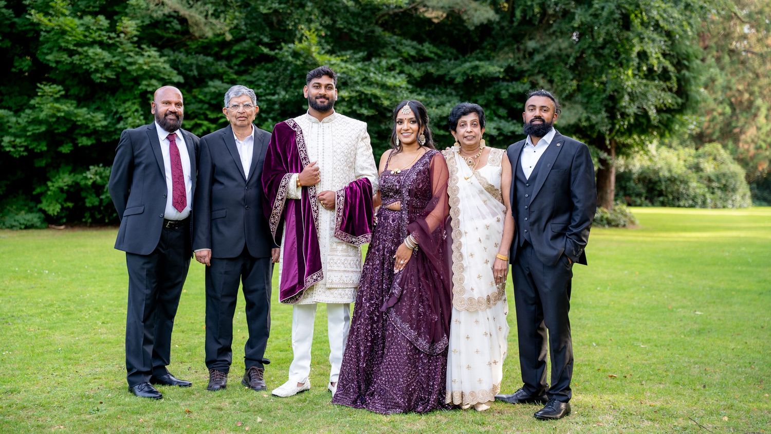 Large group of six wedding guests in traditional Indian and formal attire posing on a green lawn outdoors.