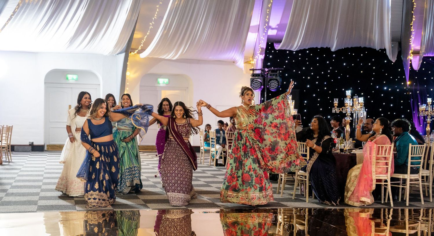 A group of women in colorful Indian attire dance joyfully together at a beautifully decorated wedding reception.