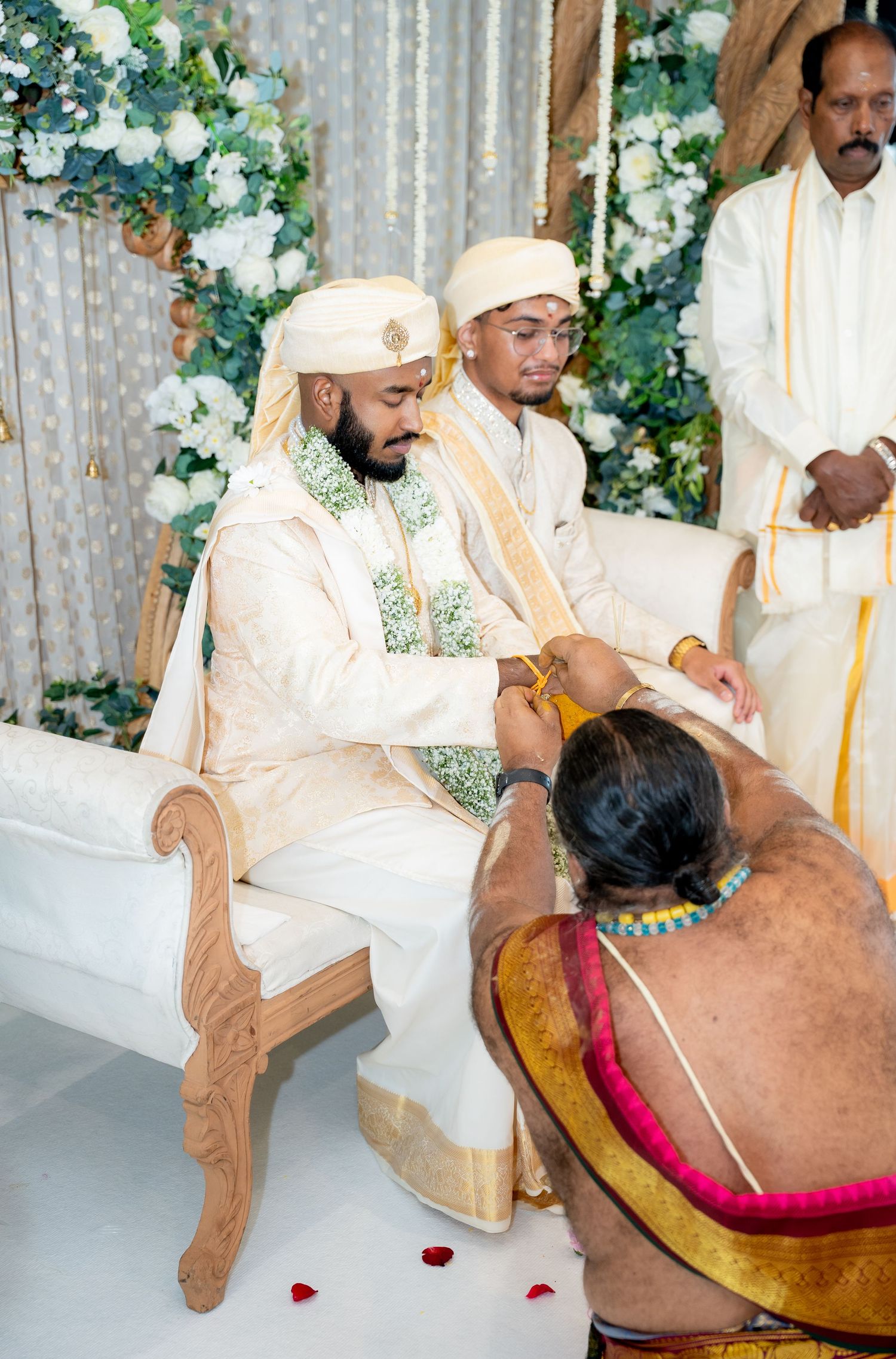 Groom in cream sherwani receiving sacred thread ritual from Hindu priest during traditional South Indian wedding ceremony.
