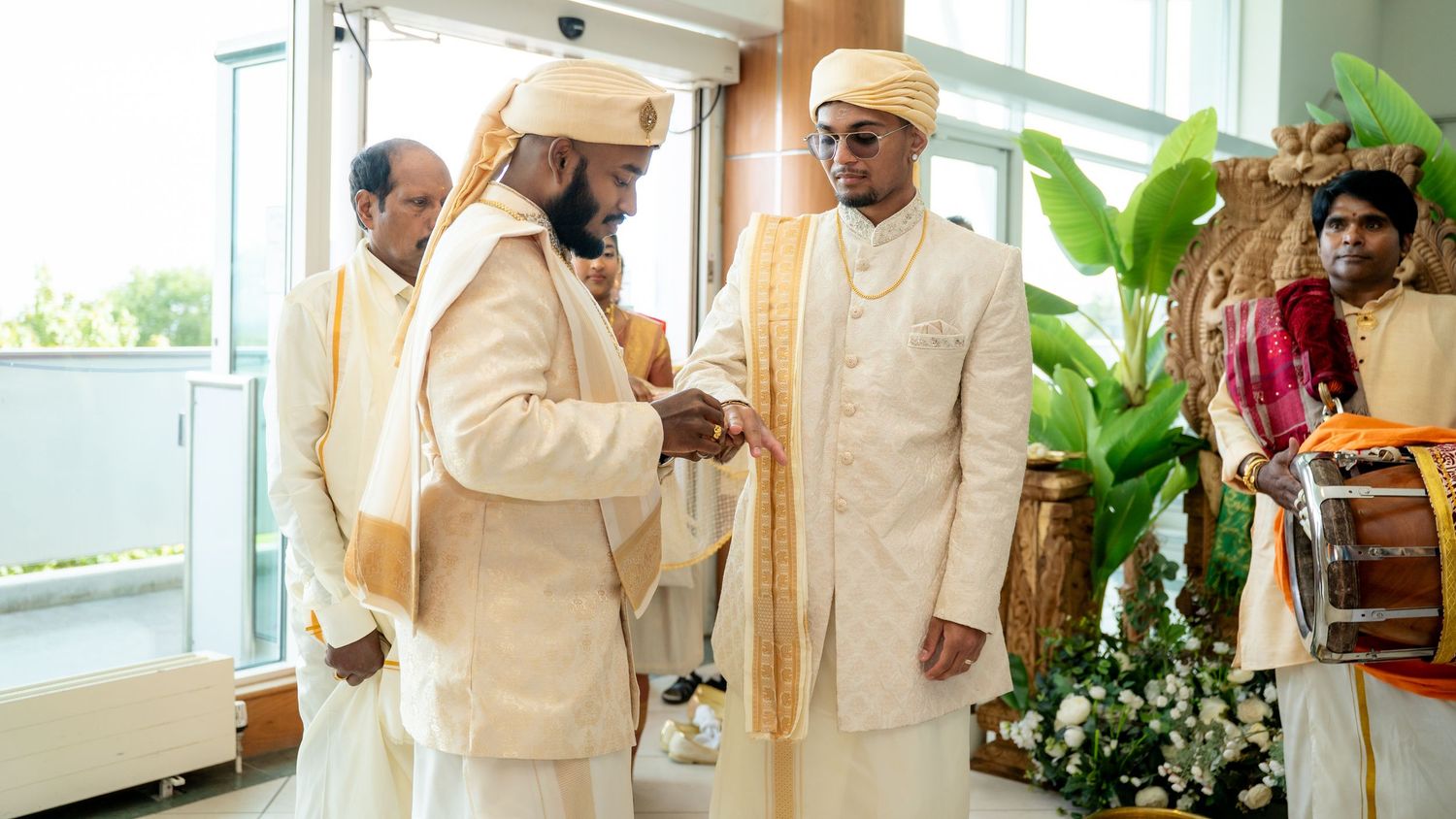 Two men in elegant cream and gold sherwanis exchanging a handshake at a traditional Hindu wedding celebration.