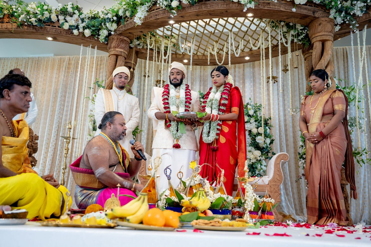 Bride and groom exchange garlands during traditional South Indian Hindu wedding ceremony with priest and guests.