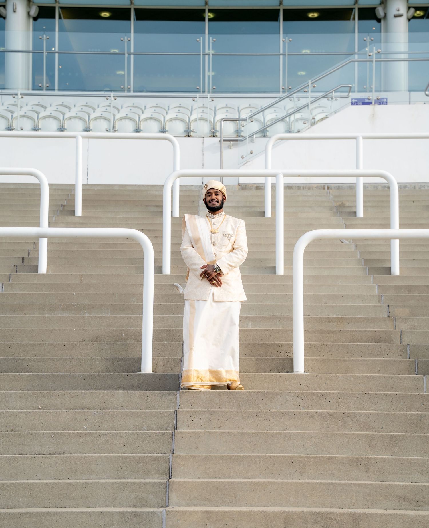 Man in white traditional wedding attire smiling on wide stadium steps with seating behind.