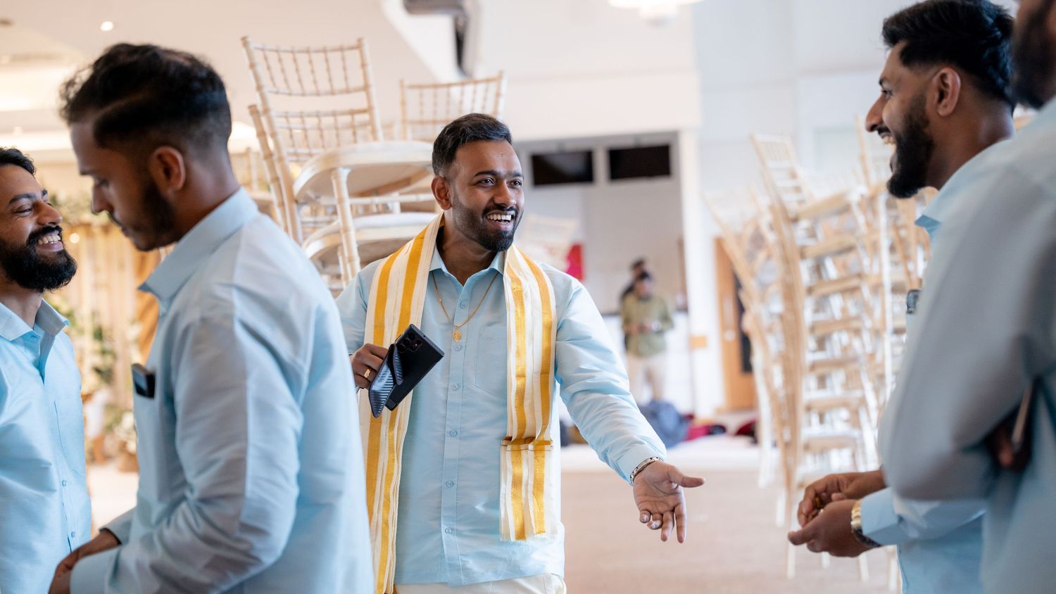 Indian groom in blue kurta and gold stole greets smiling guests at wedding venue.