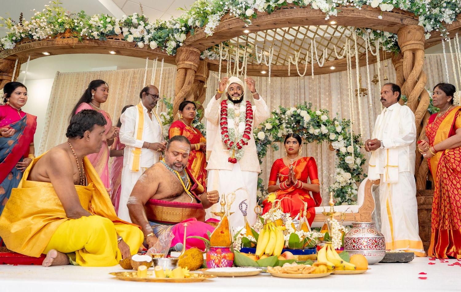 Hindu wedding rituals performed by priests as couple stands under floral mandap with family gathered around.